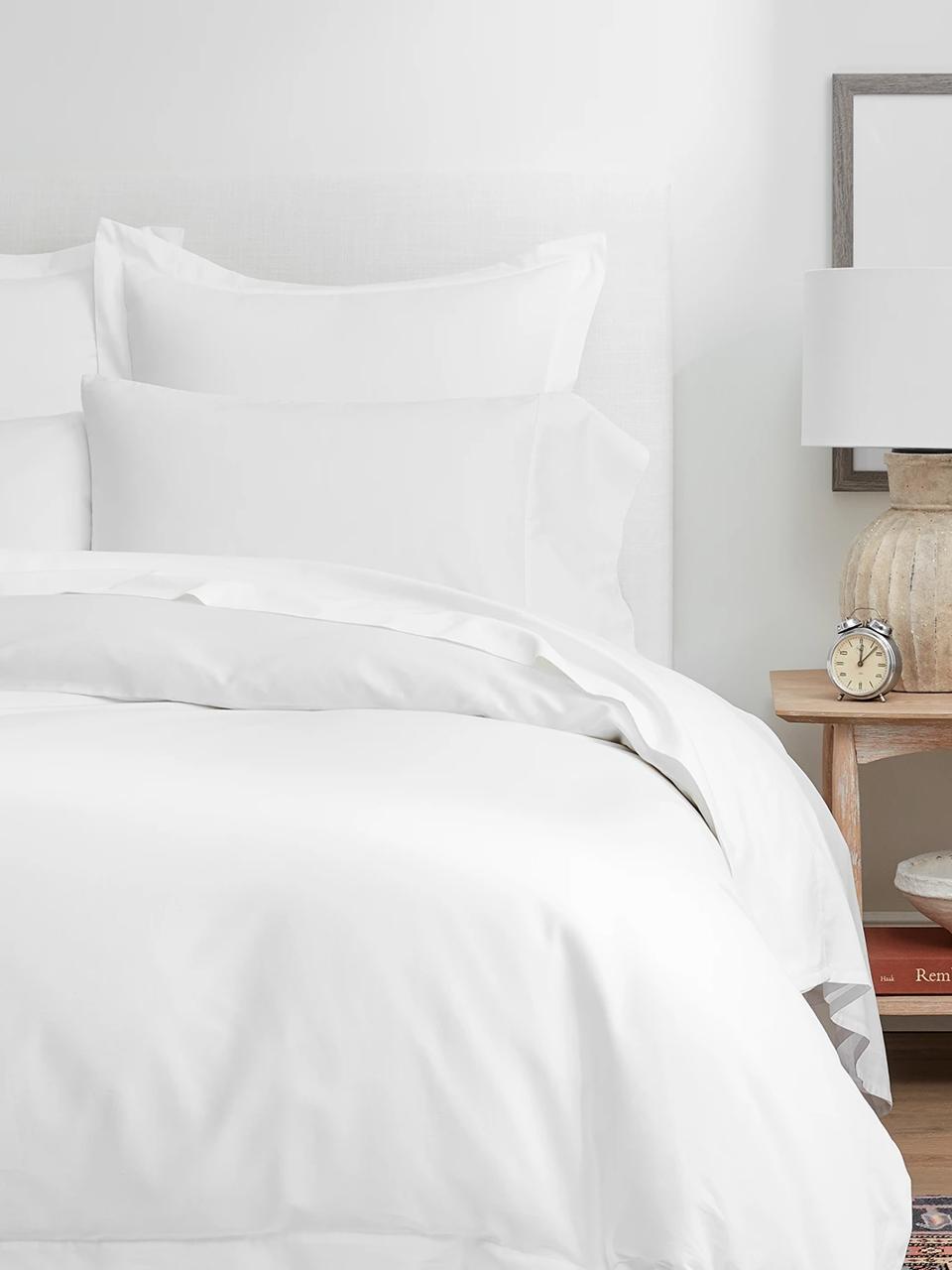 Boll & Branch Signature Hemmed Sheet Set in white displayed on a cozy bed with a side table topped with a lamp and clock. 