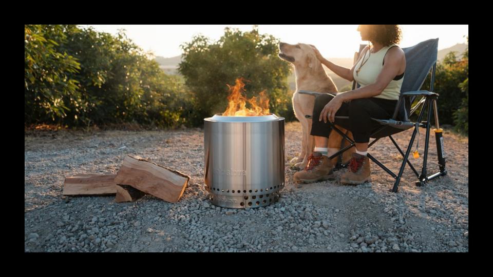 Woman sitting in a camp chair outside with her dog in front of a lit Solo Stove Bonfire. 