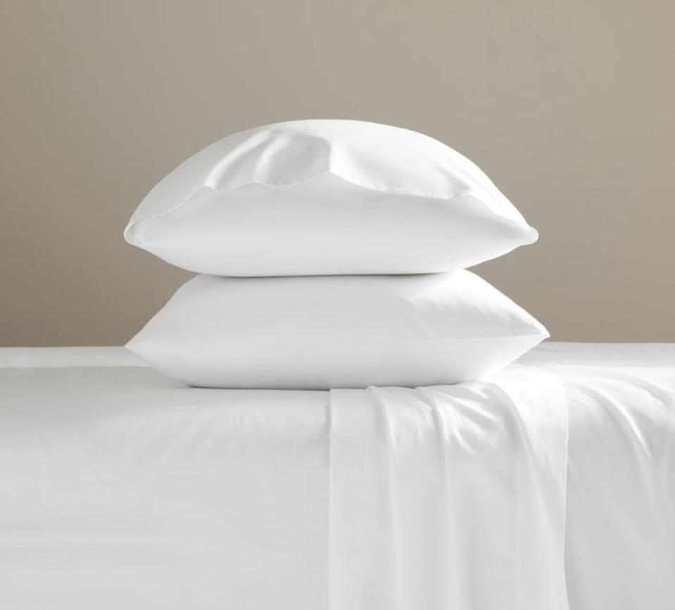 Two pillows in white pillowcases stacked on bed with white sheets against tan background.