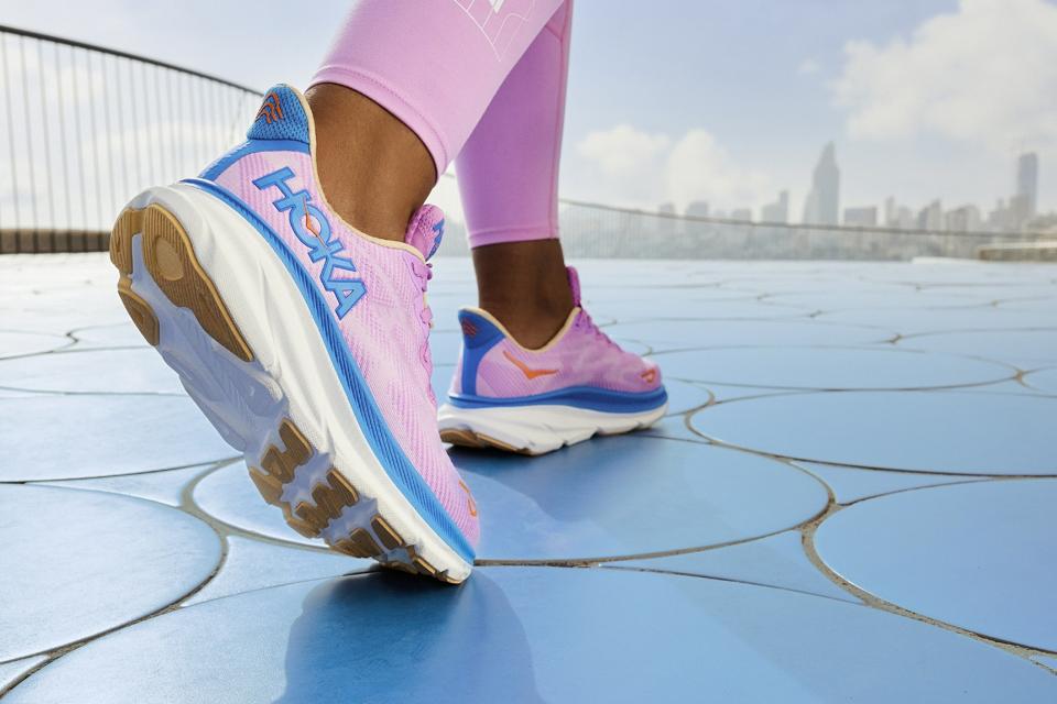 A woman wearing a pair of pink Hoka Clifton 9 shoes with pink leggings on a blue surface