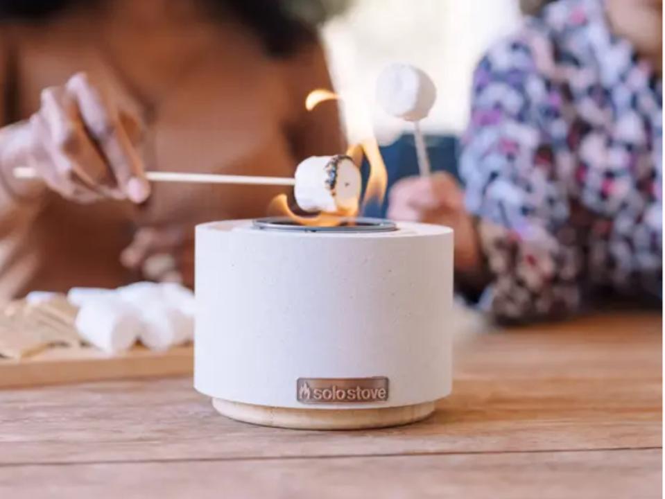 photo of white & light brown mini tabletop fire bowl with fire on wooden surface with 2 women in background toasting marshmallows over it