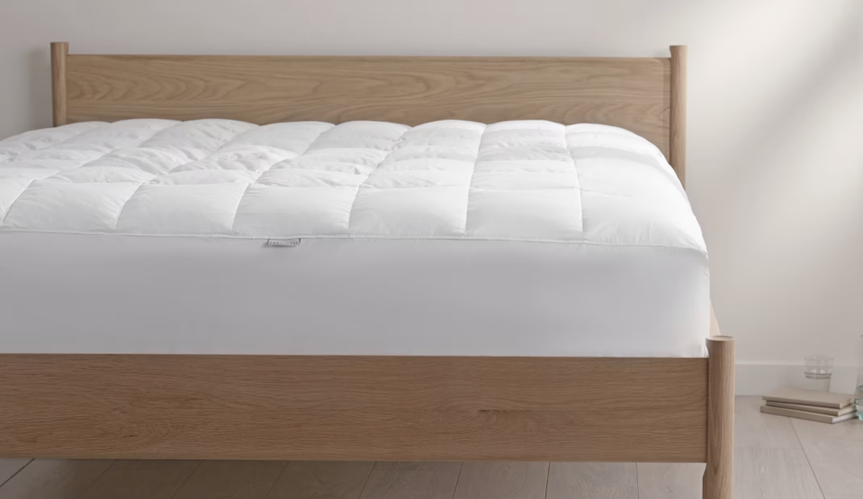 A White mattress pad on top of a mattress perched on a wooden bed frame. 