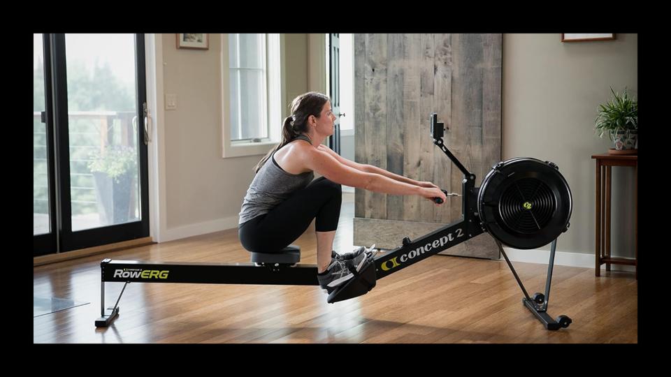 A woman using the RowErg Concept2 rowing machine in a bright room next to windows