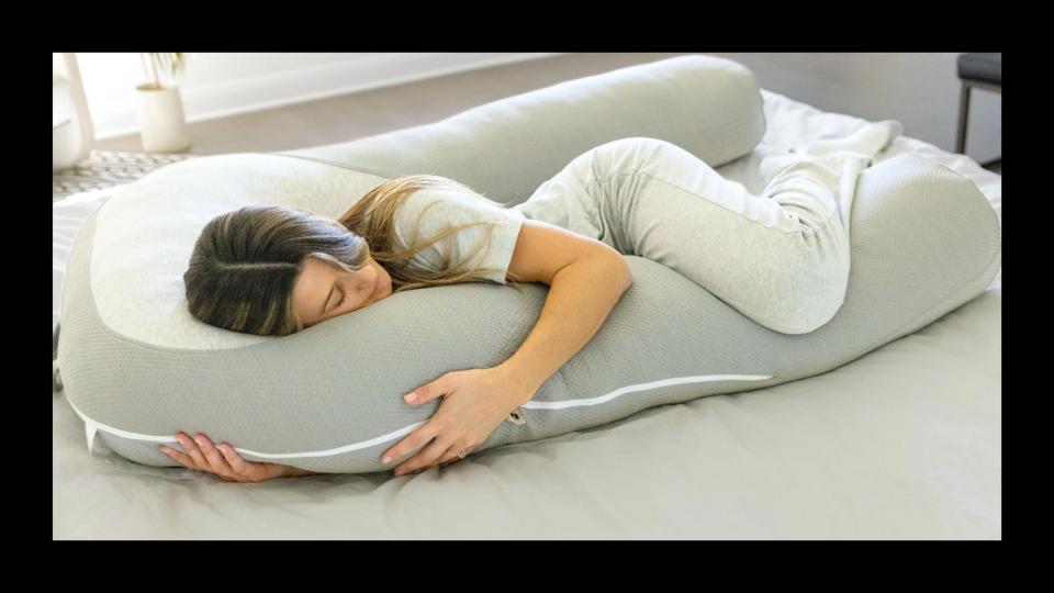 Woman cradling a gray, U-shaped body pillow on a bed.