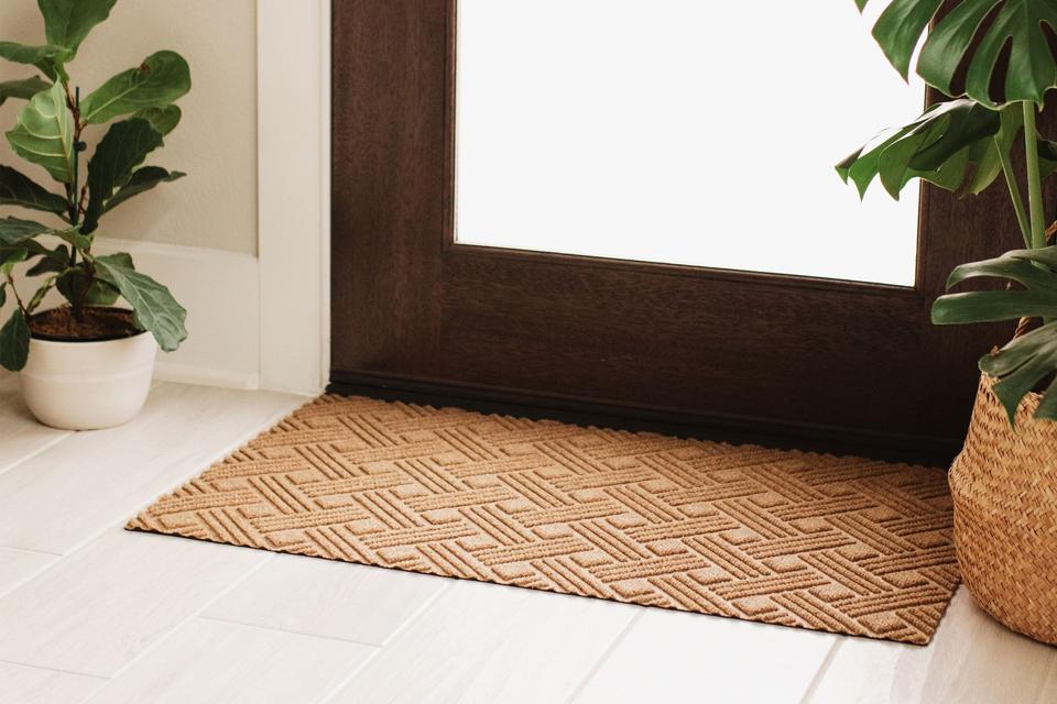 A tan Waterhog Lux Thatch Doormat indoors next to two potted plants