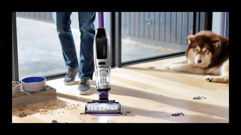 A stick vacuum cleaning up dirt on a wooden floor while a dog is featured in the background.