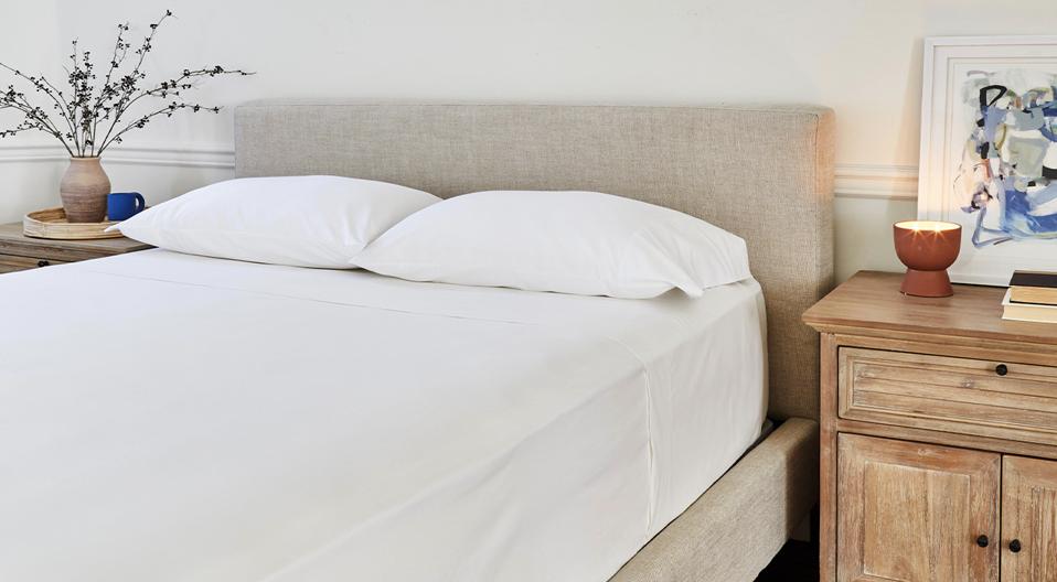 The Saatva Percale Sheets in white on a beige upholstered bed frame. 