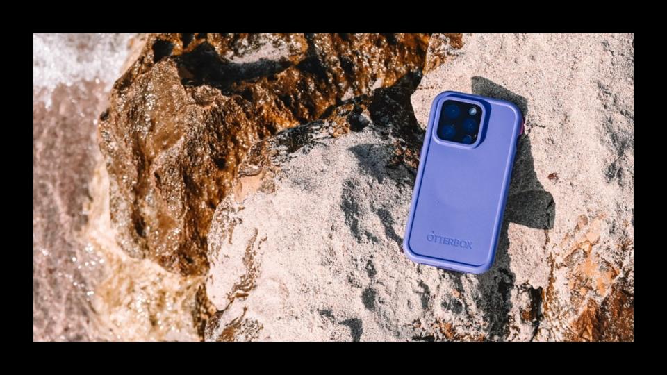 The Otterbox FRE Series Waterproof Phone Case in purple on a sandy rock next to the ocean.