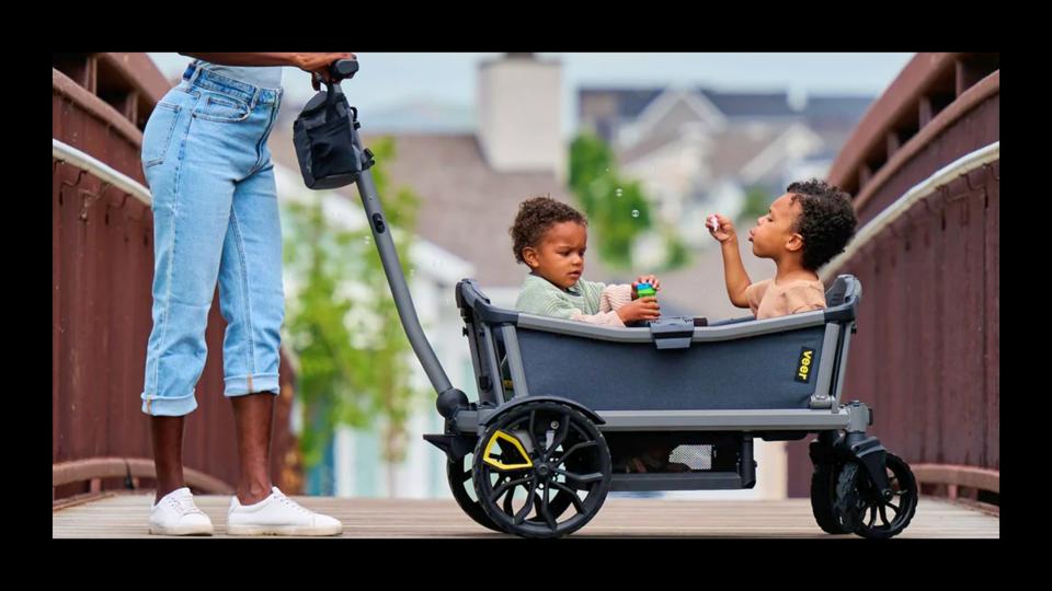 photo of gray veer cruiser city stroller wagon w/ 2 toddlers inside blowing bubbles w/ mom