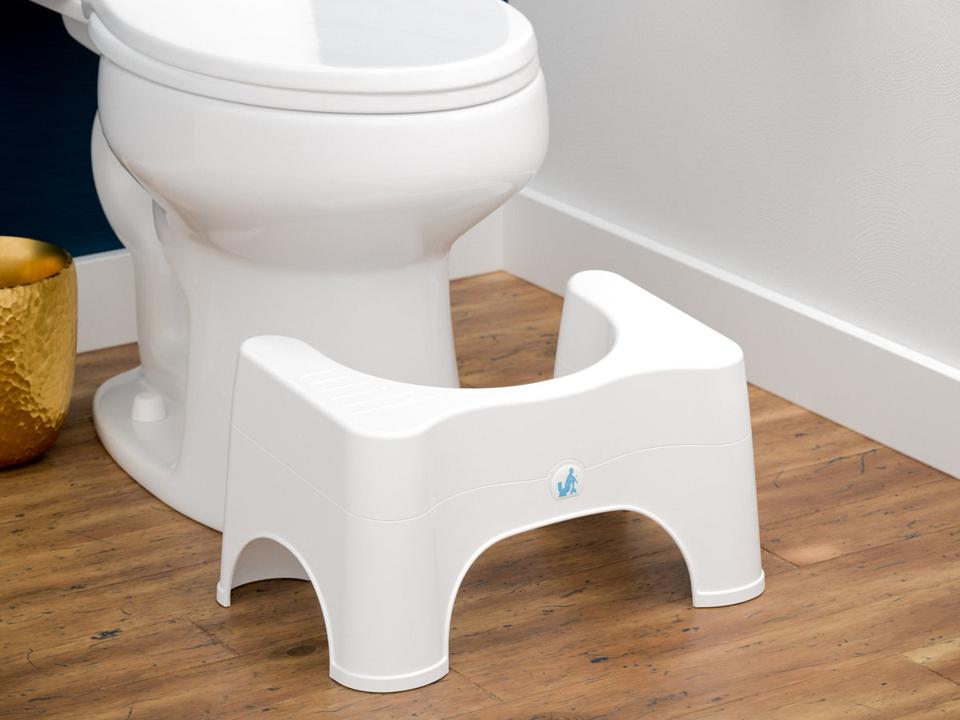 white squatty potty next to white toilet on wood floor next to white wall with gold bin