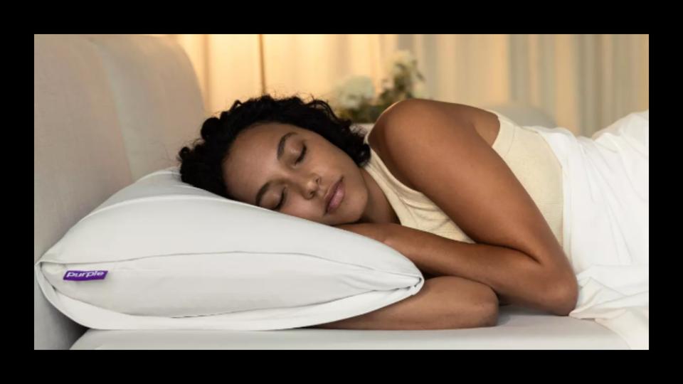 A woman sleeping on her side on the Purple Harmony Pillow facing the camera on a bed