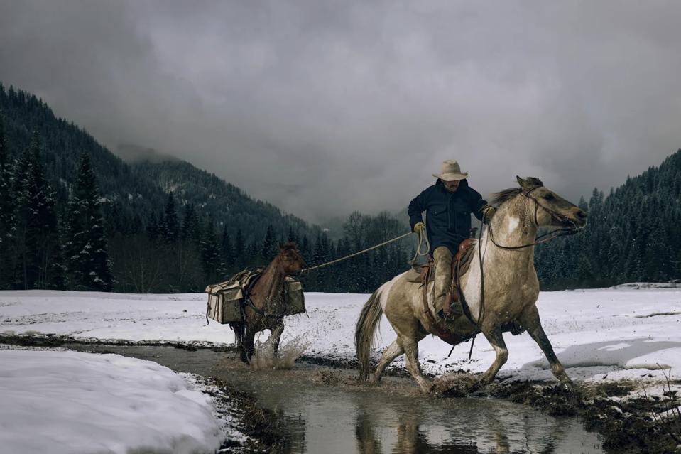 A man riding a horse through a river in the mountains with snoe on the ground