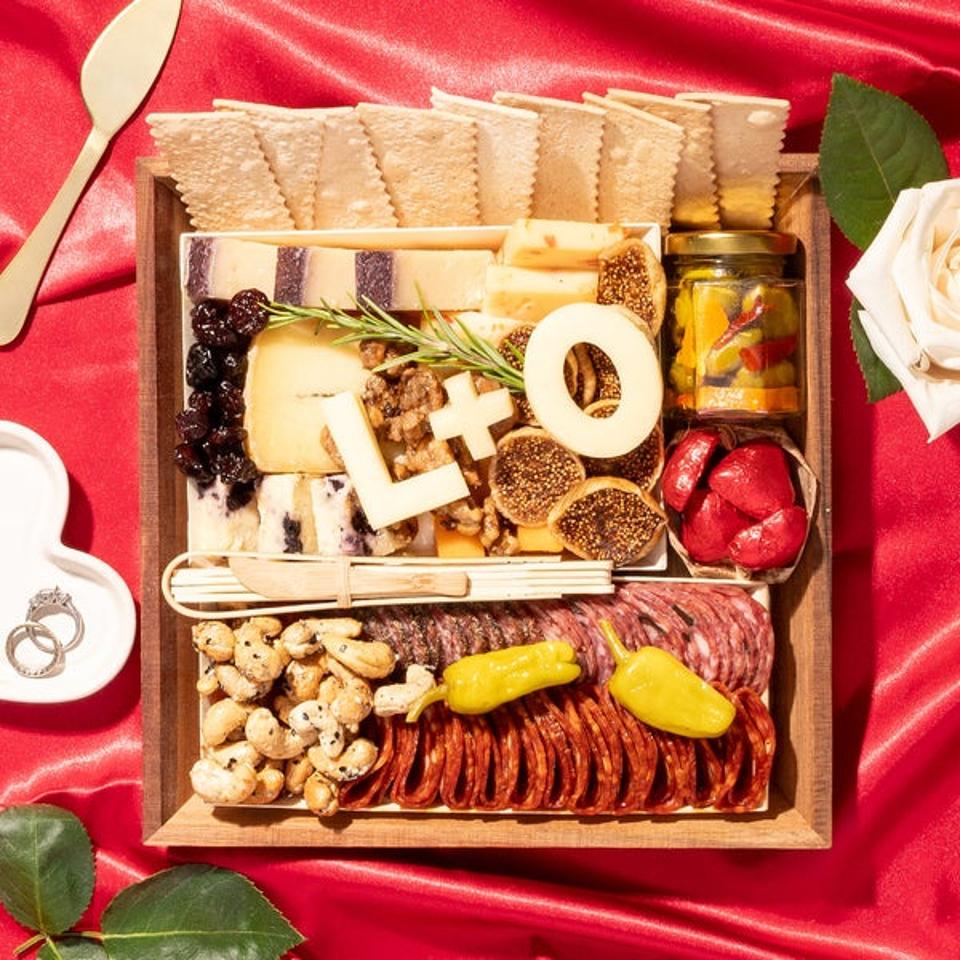 Cheese & Charcuterie Board on red tablecloth with cheese initials figs cashews pickles 