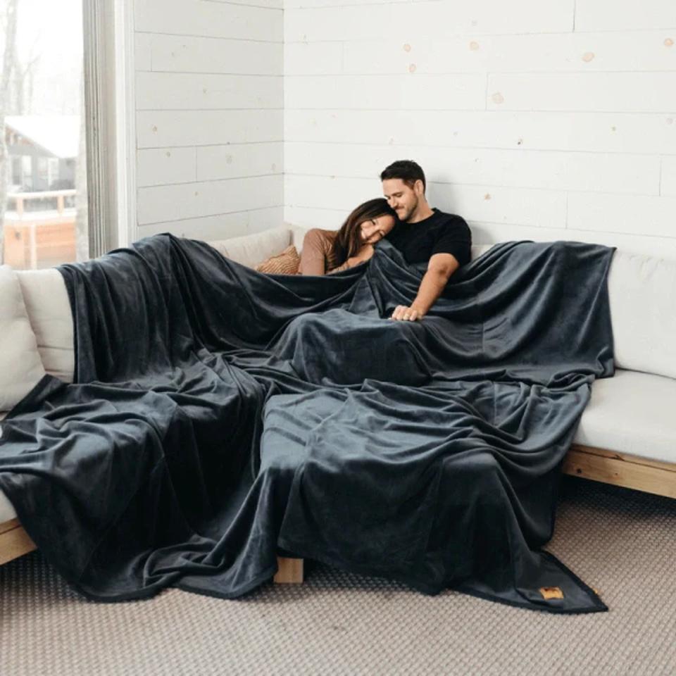 A couple using the Big Blanket Co. Original Stretch Blanket in the color Smoke on a couch 