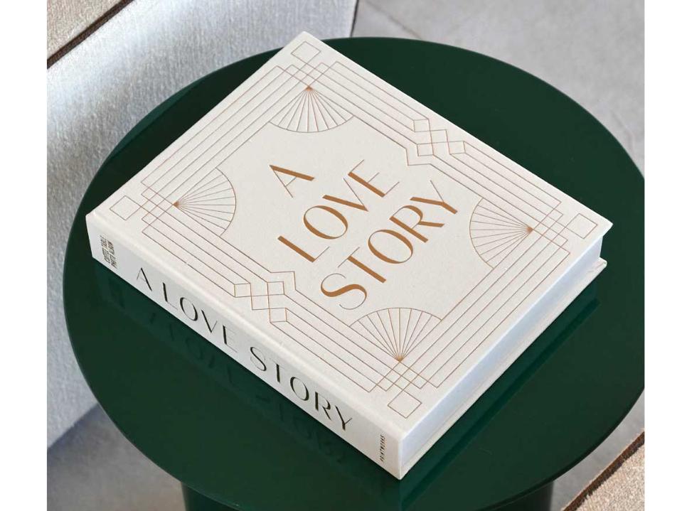 white hardcover scrapbook on green coffee table with 'a love story' in gold foil in front with offwhite wall in background