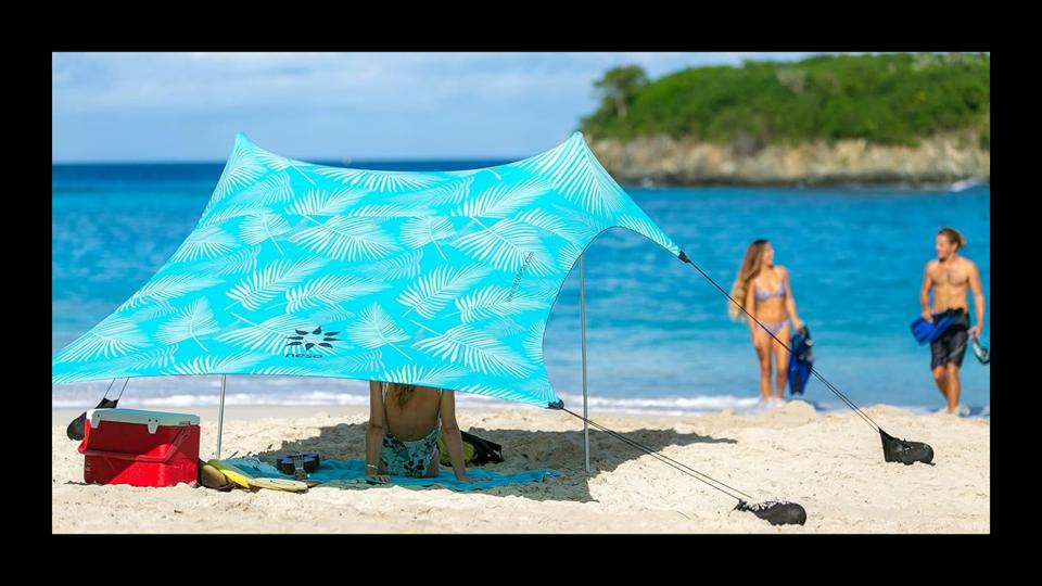 A woman sitting under a colorful beach canopy in the sand with blue waters in the distance