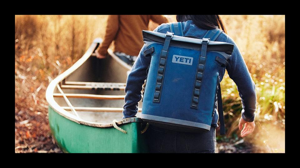 A person hauling a canoe through the brush while wearing a small backpack cooler