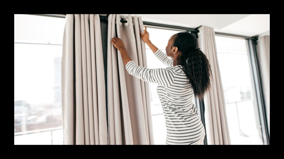 A woman in a striped shirt hanging curtains on a rod. 