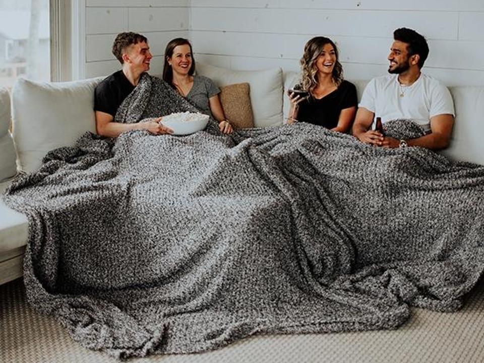 Four people sitting on a couch while using the Big Blanket Co. Premier Plush Blanket in the Black Heather color