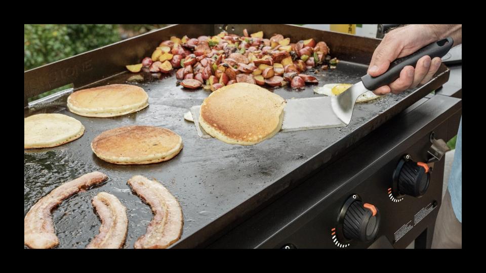 A man grilling a pancakes, bacon and potatoes on a flat-top grill. 