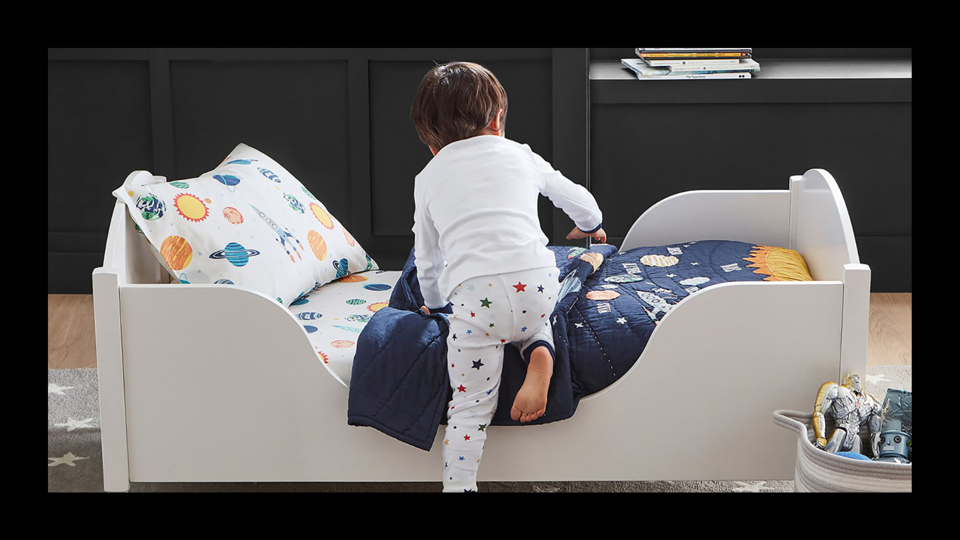 Little boy climbs into a toddler white bed with space-themed bedding.