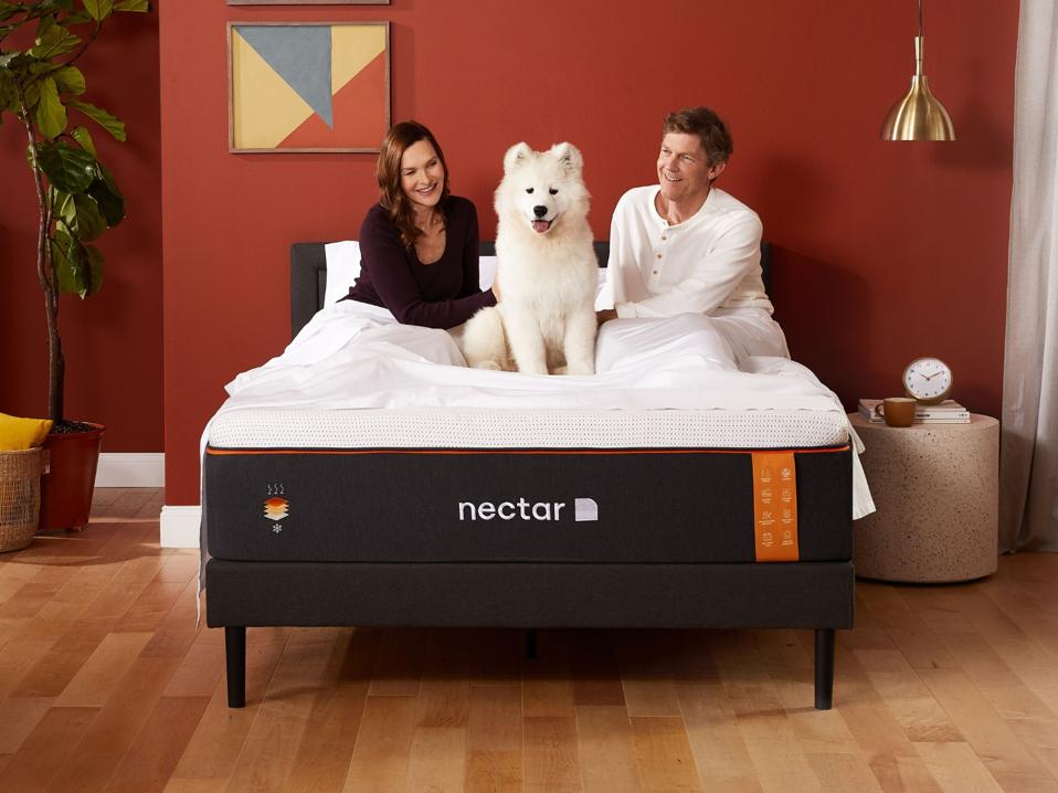 An unsheeted Nectar mattress topped with American-flag themed pillows in a well-lit room below a window.