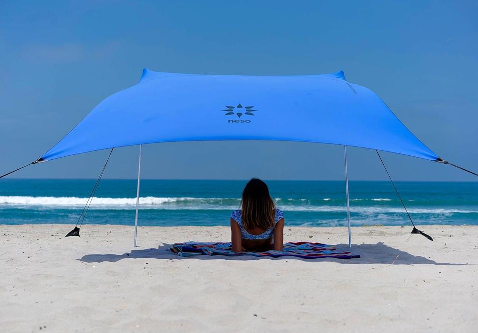 A white beach canopy in dark blue erected in the sand with a woman sitting underneath it