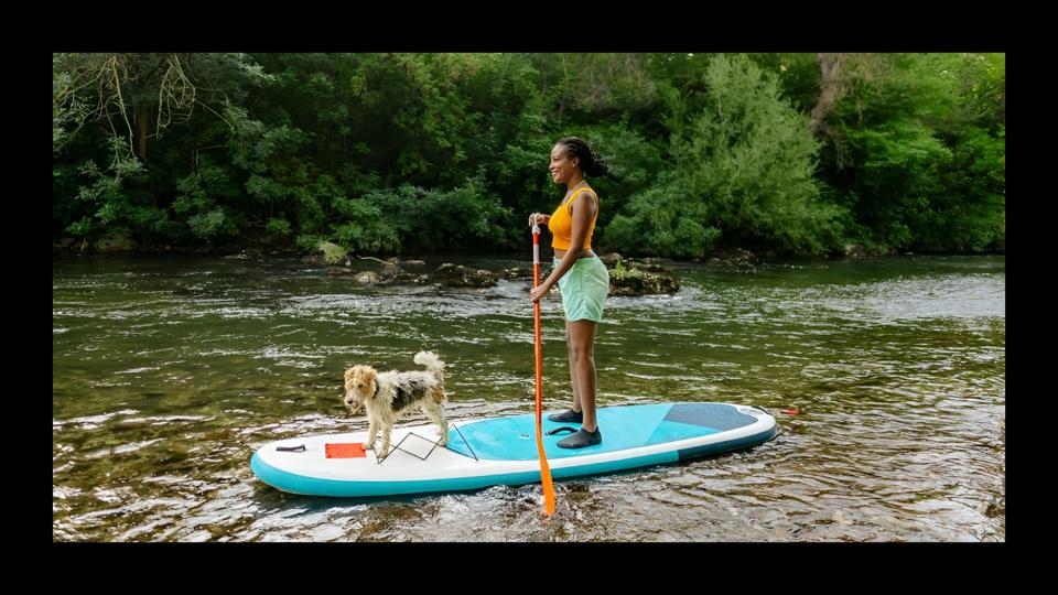 A woman maneuvering a paddle board on a river with her dog