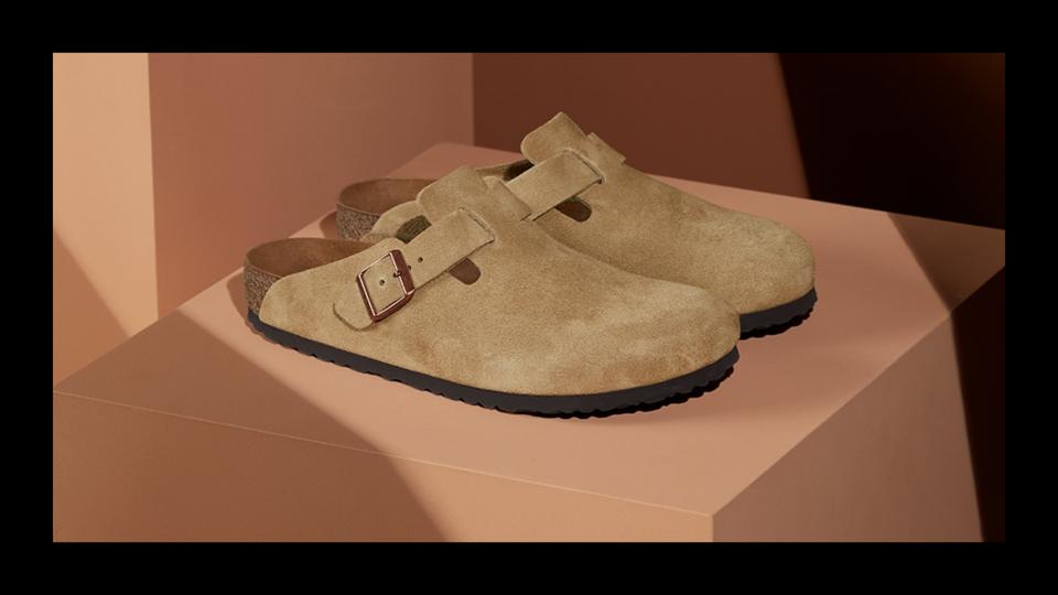 A pair of taupe suede Birkenstock Boston Clogs sitting on a peach display cube in a showroom. 