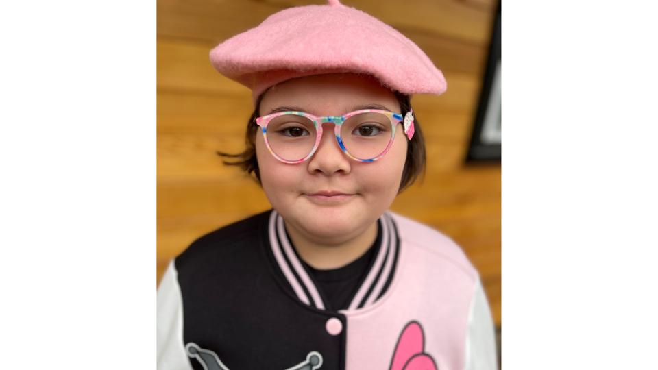 young girl with pink hat & jacket wearing pink & blue zenni optical eyeglasses against wood wall