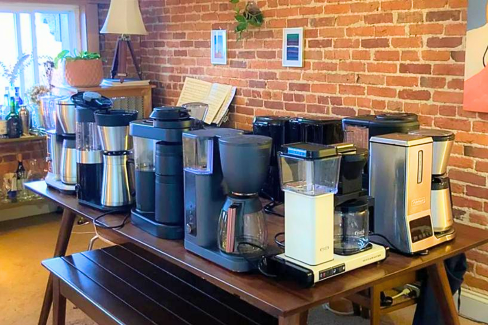 10 drip coffee makers on a brown picnic-style table in living room with brick wall and paintings