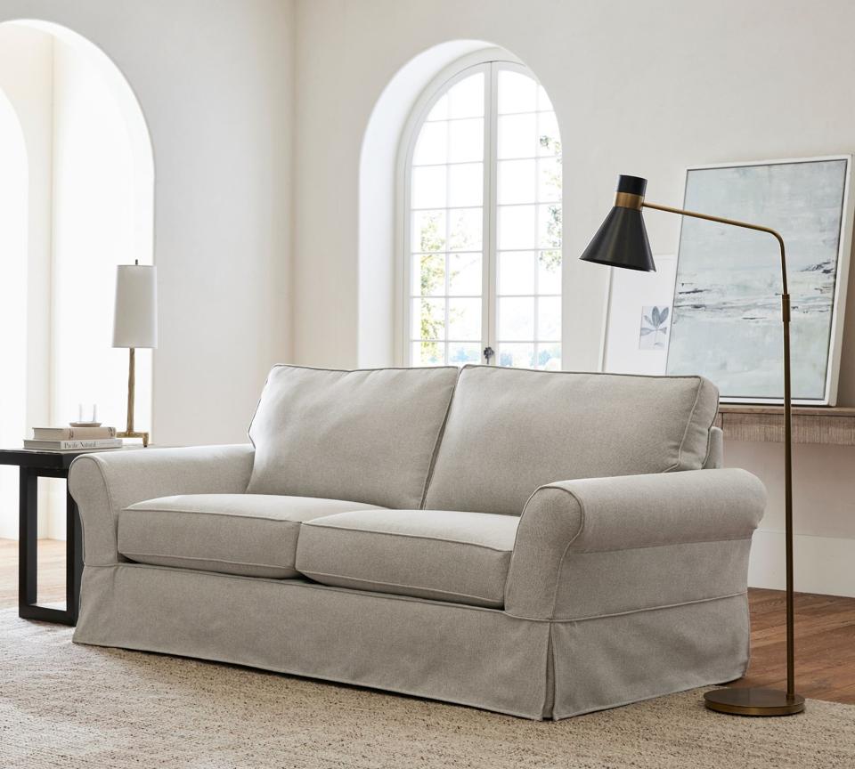 The PB Comfort Roll Arm Slipcovered Sofa in a white room with a lamp