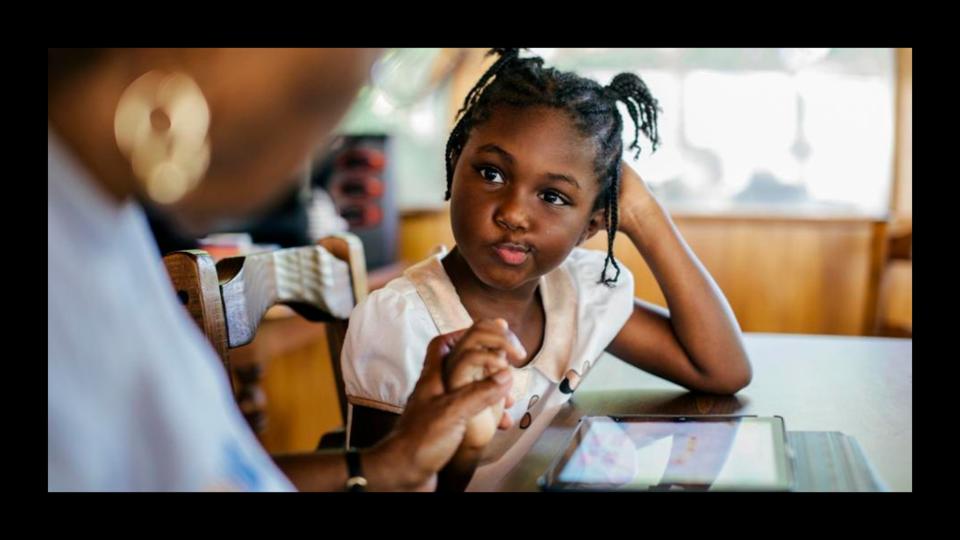 Girl sitting at an iPad on a table. She looks at her parent, who is out of focus in the foreground.