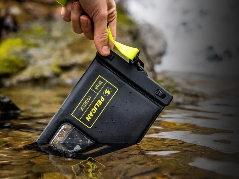 A hand dipping a phone in a Pelican Marine waterproof phone protectors into a creek