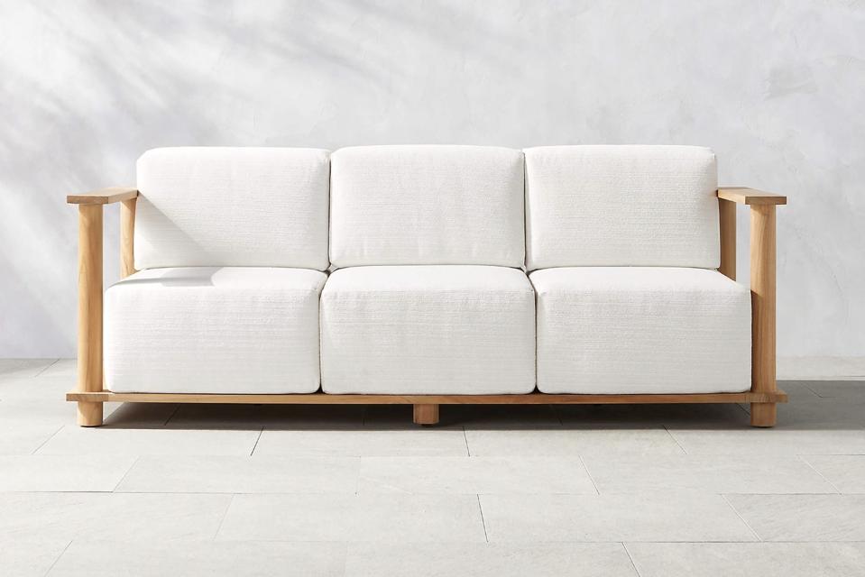 CB2 Pinet Teak Outdoor Sofa (84-Inch) on a stone patio in front of a white wall