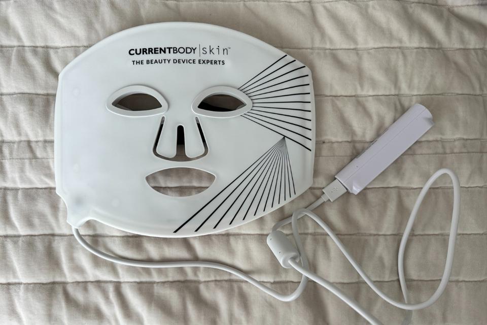 CurrentBody Skin LED Light Therapy Face Mask and controller against white bedspread