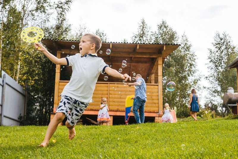 Un petit garçon court en soufflant des bulles de savon. Arrière-cour, famille en arrière-plan.