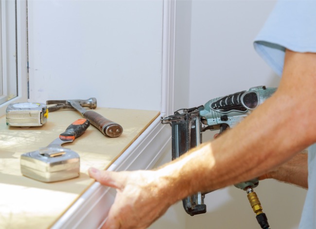 Un homme utilise une cloueuse de finition pour clouer des garnitures autour d'une fenêtre avec un assortiment d'outils à proximité.