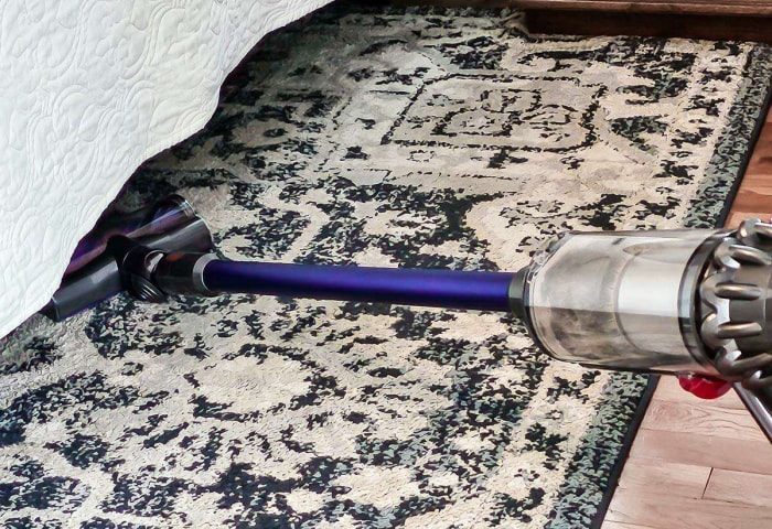 Une personne passe l'aspirateur sous un tapis noir et blanc sous un lit avec l'aspirateur-balai Dyson V15 Detect.