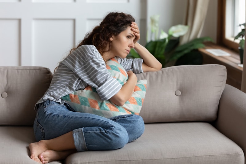 Une jeune femme frustrée assise sur un canapé, blottie contre un oreiller, regarde ailleurs par la fenêtre. Perdue dans ses pensées, une femme millénaire stressée et malheureuse regrette sa mauvaise décision, passe du temps seule dans le salon.