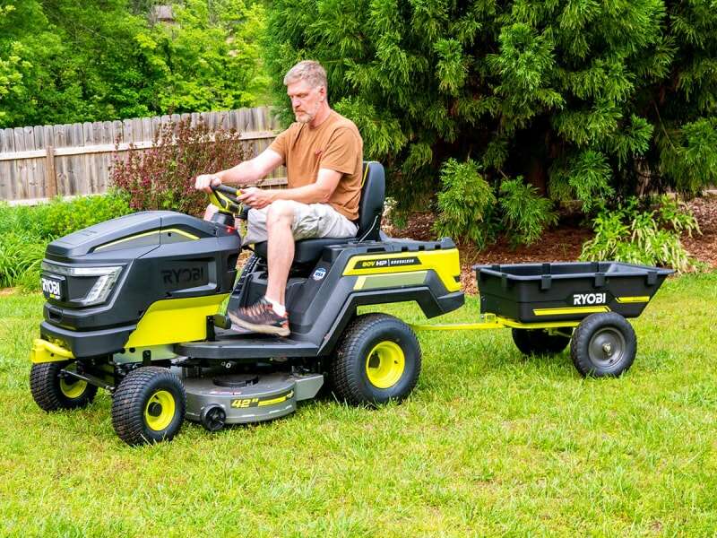 Personne en chemise marron conduisant un tracteur à gazon Ryobi 80V HP Electric sur l'herbe devant des arbres.