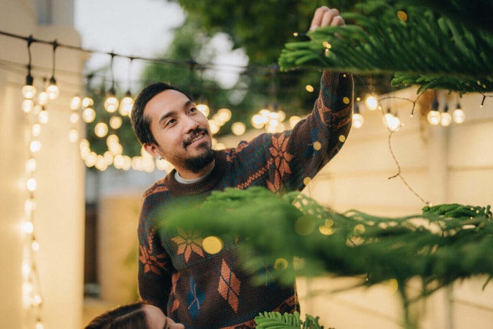 Un homme portant un pull de Noël accroche des lumières de Noël dans son jardin.