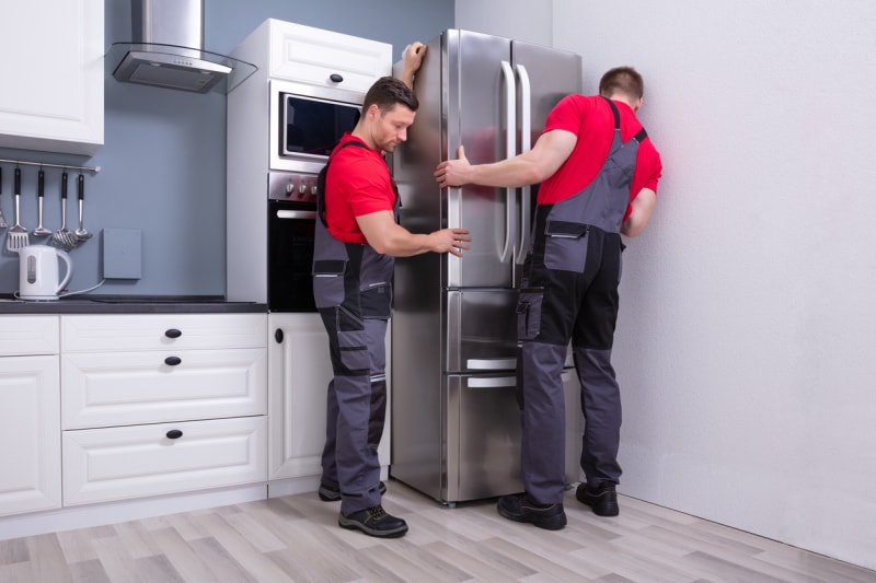 Deux jeunes déménageurs en uniforme placent un réfrigérateur moderne en acier dans la cuisine