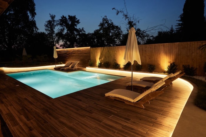 Terrasse de piscine en bois moderne avec piscine éclairée, mobilier de terrasse et parasol.