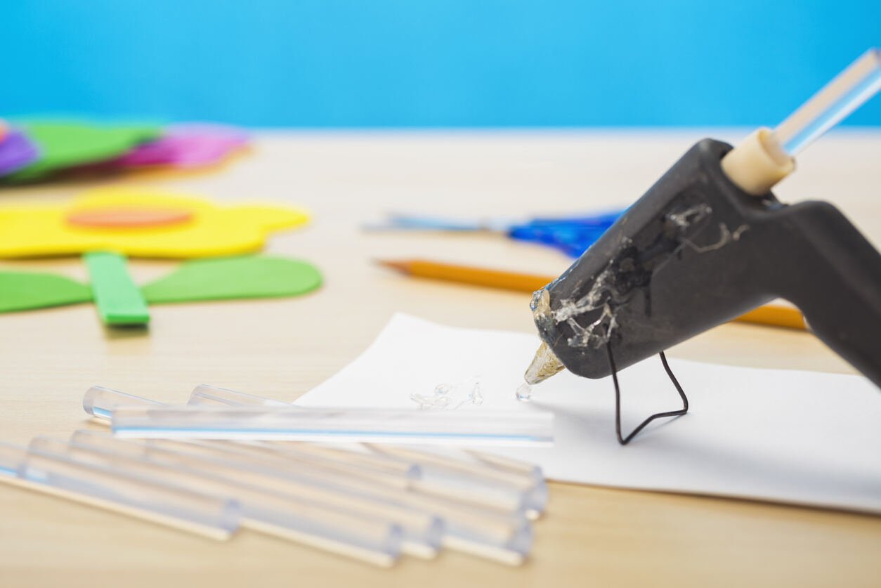 Un mini pistolet à colle chaude avec une goutte de colle transparente sortant de la pointe sur un morceau de papier blanc, avec des ciseaux et un crayon, ainsi qu'une fleur jaune créée à partir de feuilles de mousse sur une table en bois clair et un mur bleu en arrière-plan.