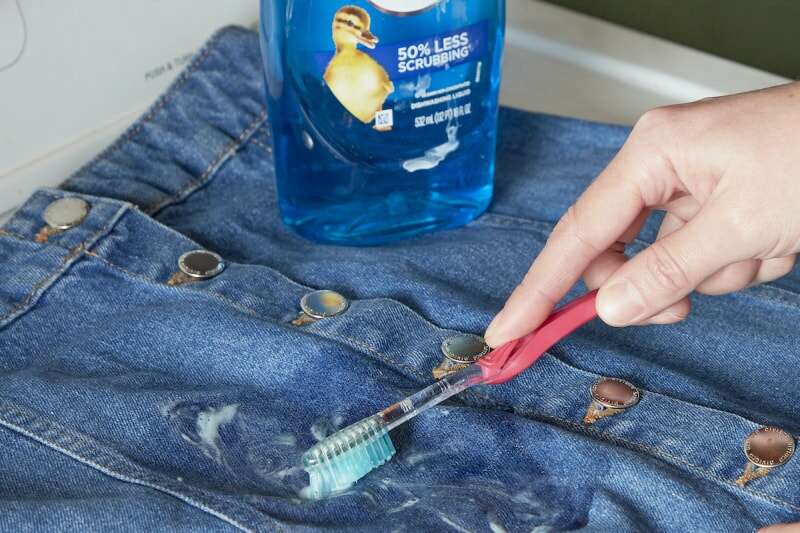 Une femme utilise une brosse à dents pour frotter du liquide vaisselle sur une tache sur une jupe en jean.