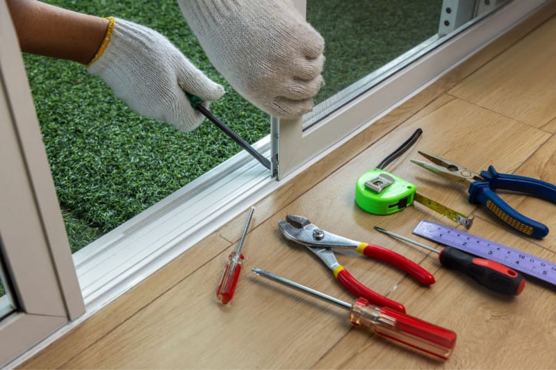 Les mains d'une personne réparent la moustiquaire d'une porte-fenêtre coulissante à l'aide d'un tournevis à tête plate. Un ensemble d'outils se trouve à côté de la porte.