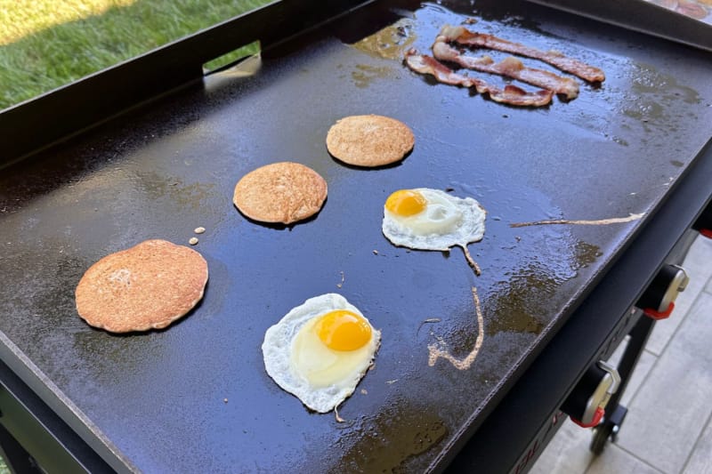 Trois crêpes, deux œufs et trois tranches de bacon cuisent sur une plaque chauffante.