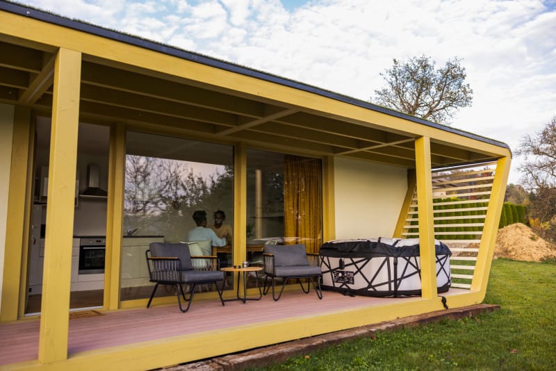 Une petite maison trapézoïdale en forme de conteneur est dotée de grandes fenêtres donnant sur un petit patio entièrement meublé et doté d'un jacuzzi.