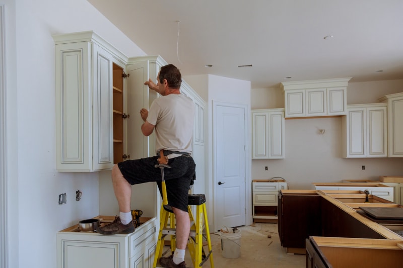 Un homme en vêtements de travail installe des armoires de cuisine blanches dans une cuisine nouvellement construite.